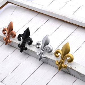 May include: Four fleur-de-lis decorative accents in copper, black, silver, and gold. The accents are arranged diagonally on a white wooden surface, showcasing their intricate design and metallic finishes.