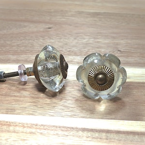 May include: Two clear glass flower-shaped cabinet knobs with brass accents. One knob is fully assembled with a screw and nut, while the other is shown from the front. The knobs are on a wooden surface.