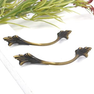 May include: Two antique bronze-colored drawer pulls with a curved handle design. Each pull features decorative leaf-shaped accents on both ends. The hardware is set against a white background with greenery in the upper corners.