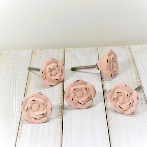 May include: Five rose-shaped cabinet knobs in a soft peach color with black accents. The knobs are made of ceramic and have a metal screw for installation. The knobs are arranged on a white wooden surface.