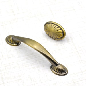 May include: An antique brass-finished cabinet pull and matching knob. The pull is a curved handle with decorative ends, and the knob is oval-shaped with a floral design. These hardware pieces are likely for furniture or cabinetry.