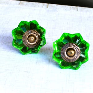 May include: Two green glass cabinet knobs with a flower-like design and bronze-colored metal accents. The knobs are set against a white, textured surface. Each knob has a diameter of approximately 1.5 inches.
