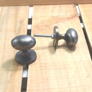 May include: Two antique-style cabinet knobs with an oval shape and a dark, metallic finish. One knob is assembled with a screw and washer, while the other is separate. The knobs are on a light-colored wooden surface.