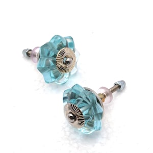 May include: Two decorative cabinet knobs. Each knob features a faceted, light blue glass flower design with a silver-colored metal base and a threaded post for installation. The knobs are set against a white background.