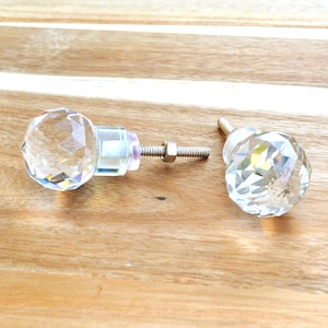 May include: Two clear crystal cabinet knobs with faceted surfaces. One knob is attached to a metal screw with a nut, while the other is separate. The knobs are on a light brown wooden surface.