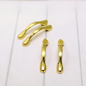 May include: Four gold-colored cabinet handles. Two are curved with a leaf design at each end, and two are straight with a twisted design. The handles are made of metal and have a shiny finish.