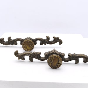 May include: Two antique bronze-toned drawer pulls with ornate, scrolling designs. Each pull features a central, circular knob with a raised design. The hardware is likely for furniture restoration.