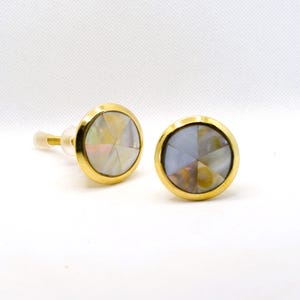 May include: A pair of round stud earrings with a gold-colored bezel. The earrings feature a mother-of-pearl center with a geometric design. One earring is slightly angled, showing the post.