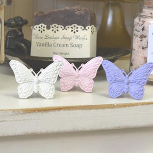 May include: Three decorative butterfly ornaments in white, pink, and purple. The butterflies are made of metal and have a detailed design. They are displayed on a white shelf.