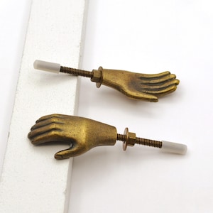 May include: Two antique brass-toned hand-shaped cabinet knobs. Each knob features a detailed hand design with a threaded rod, nut, and a white plastic tip. The knobs are set against a white background.