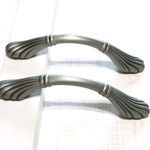 May include: Two silver-colored cabinet handles. Each handle has a curved bar with decorative ends featuring a fan-like design. The handles are made of metal and are set against a white background.