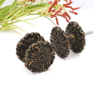 May include: Close-up of four antique bronze cabinet knobs with a floral design. The knobs have a detailed, ornate pattern and a screw for installation. Decorative hardware for furniture or home decor.