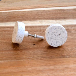 May include: Two white cabinet knobs with a speckled design. The knobs are round and have a metal screw and washer for installation. The knobs are on a wooden surface.