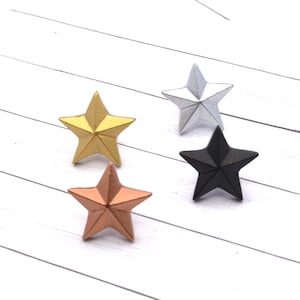 May include: Four star-shaped decorative objects in gold, silver, copper, and black. The stars are arranged on a white, wood-grain surface. The objects are likely used for crafts or home decor.