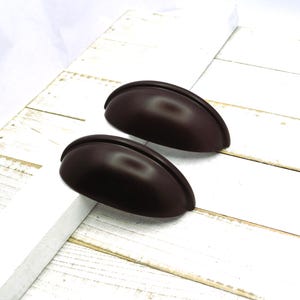 May include: Two dark brown, cup-style cabinet pulls. The pulls are semi-circular with a smooth, matte finish. They are set against a white, weathered wood background, suggesting a rustic or farmhouse aesthetic.