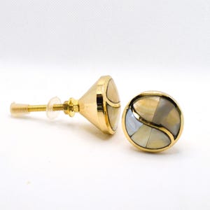 May include: Two gold-toned cabinet knobs with a mother-of-pearl inlay. One knob is cone-shaped, the other is round. The knobs have a gold screw and a clear plastic washer. The knobs are for furniture.