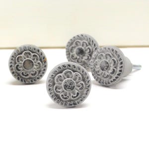 May include: Four gray cabinet knobs with a distressed finish. Each knob features a central floral design surrounded by a decorative border. The knobs are made of a solid material and have a metal screw for attachment. Ideal for adding a vintage touch to furniture.