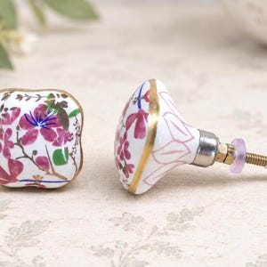 May include: Two square ceramic cabinet knobs featuring a white base with a painted floral pattern in shades of pink, green, and blue. Each knob has a gold-toned metal rim and a threaded metal bolt with a nut and a small translucent washer.