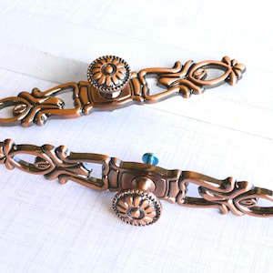 May include: Two ornate bronze-colored cabinet pulls with floral and scroll designs. Each pull has a central, round knob with a flower-like pattern and a blue accent. The pulls are approximately 6 inches long.