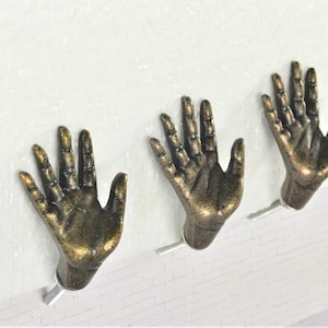 May include: Three bronze-colored hand-shaped wall hooks. Each hook is mounted on a white surface. The hooks are detailed with visible knuckles and fingernails, adding a unique touch to any space.
