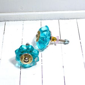 May include: Two turquoise glass cabinet knobs with a faceted design. Each knob has a gold-colored metal base and a silver screw. The knobs are displayed on a white wooden surface.