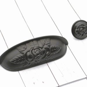 May include: Black cabinet hardware set featuring a rose design. The set includes a cup pull and a round knob. The hardware is made of metal and has a matte finish. The rose design is embossed on both pieces.