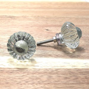 May include: Two clear glass cabinet knobs with a fluted design and a silver-toned metal base. The knobs are on a wooden surface, showcasing their reflective qualities. These decorative knobs are ideal for furniture upgrades.
