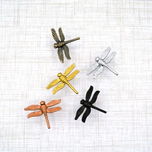 May include: Five dragonfly-shaped decorative hardware pieces in various metallic finishes: bronze, gold, silver, copper, and black. The dragonflies are arranged on a light-colored, textured surface. These decorative accents could be used for cabinet pulls or drawer knobs.