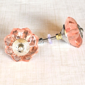 May include: Two decorative cabinet knobs with a flower-shaped design. The knobs are made of pink glass with a faceted surface and metallic hardware. The hardware includes a screw, nut, and washer. The knobs are designed for furniture.