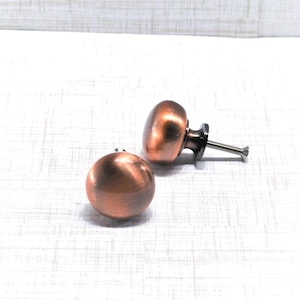 May include: Two copper-colored cabinet knobs with a rounded shape. One knob is lying on its side, while the other is upright, showing the screw. The knobs have a metallic finish and are designed for furniture.