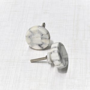 May include: Two white and gray marble cabinet knobs with silver hardware. One knob is round, and the other is a flattened hexagon shape. The knobs are set against a white background.