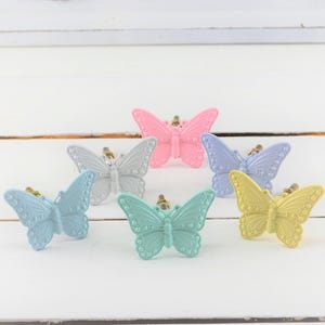 May include: A collection of six butterfly-shaped cabinet knobs in pastel colors: light blue, gray, pink, lavender, mint green, and yellow. Each knob has a gold-colored stem. The knobs are arranged on a white wooden surface.
