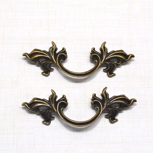May include: Two antique bronze-toned drawer pulls with a decorative, ornate design. Each pull features a curved handle and stylized floral accents. The hardware is designed for furniture and cabinetry.
