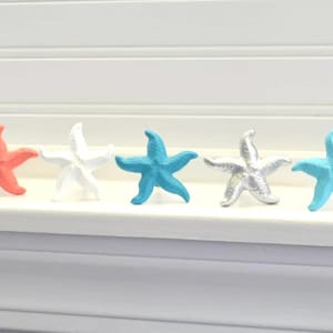 May include: A row of six starfish decorations in various colors: teal, coral, white, blue, silver, and light blue. The starfish are arranged on a white surface, likely a windowsill, with a white shutter background.