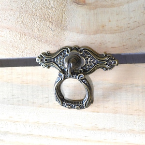 May include: Close-up of an antique brass drawer pull with an ornate design. The pull features a decorative backplate and a ring handle. The hardware is mounted on a light-colored wooden surface, showcasing the intricate details of the metalwork.