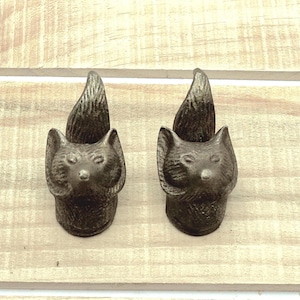 May include: Two bronze-colored cabinet knobs, each shaped like a fox head with a bushy tail. The knobs have a detailed, textured surface and are set against a light wood background. These decorative knobs are ideal for adding a rustic touch to furniture.