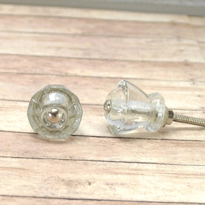May include: Two clear glass cabinet knobs with a faceted design and a silver-colored metal screw. One knob is upright, and the other is lying on its side. The knobs are on a light brown wooden surface.