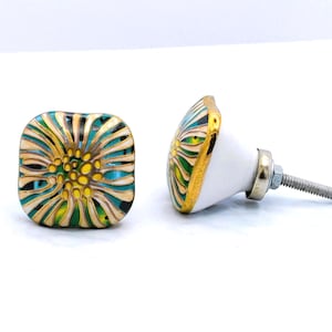 May include: Two square cabinet knobs with a floral design. The knobs feature a white base with a gold trim and a colorful floral pattern in shades of yellow, teal, and green. The knobs have a metal screw for installation.