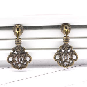 May include: Two antique bronze-colored key-shaped drawer pulls. Each pull features an ornate, openwork design with a decorative top and a key-shaped bottom. The pulls are mounted on a white surface.
