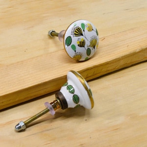 May include: Two decorative cabinet knobs with floral designs. The knobs have a white base with green leaves and yellow flowers. The edges are trimmed with gold. The hardware includes a threaded rod, nut, and washer.
