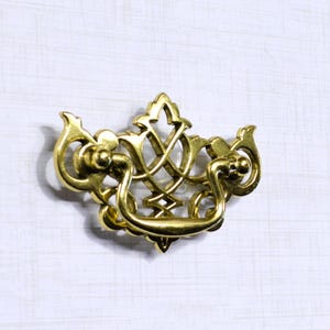 May include: A gold-colored, ornate drawer pull with a decorative design. The pull features a curved handle and intricate detailing, suitable for furniture restoration or adding a vintage touch to cabinets and drawers.