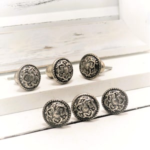 May include: Six antique-style cabinet knobs with a silver finish. Each knob features a detailed floral design within a circular frame. The knobs are arranged on a white wooden surface, suggesting a vintage or rustic aesthetic.