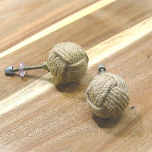 May include: Two decorative rope ball cabinet knobs with metal hardware. The knobs are made of tightly wound natural-colored rope, and are mounted on a wooden surface. The hardware includes a bolt, washer, and nut.