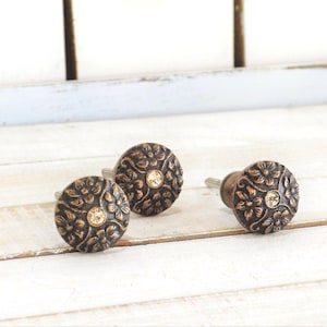May include: Three bronze-colored cabinet knobs with a floral design and a central crystal. The knobs are round and have a vintage aesthetic. They are displayed on a white wooden surface.