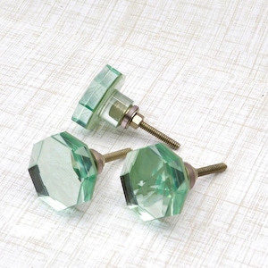 May include: Three decorative glass cabinet knobs with faceted, light green crystal-like designs. The knobs have metal hardware with threaded rods for installation. The knobs are arranged on a white, textured surface.