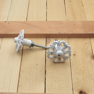 May include: White decorative cabinet knobs with a floral design and black accents. The knobs are attached to a metal screw and are set against a light wood background. The knobs are likely for furniture.