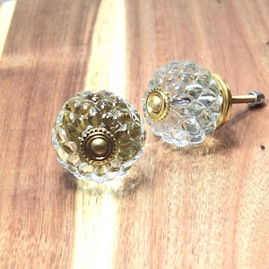 May include: Two clear glass cabinet knobs with a faceted design and brass-colored accents. The knobs have a round shape and are set against a wooden surface. One knob has a screw attached.