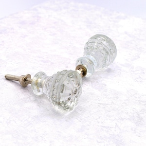 May include: Two clear glass cabinet knobs with a textured, bubble-like design. Each knob has a metal screw and washer for installation. The knobs are lying on a white surface.