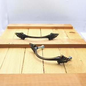 May include: Two antique bronze-colored cabinet handles with decorative leaf accents. The handles are curved and include mounting screws. They are displayed on a light-colored wooden surface.