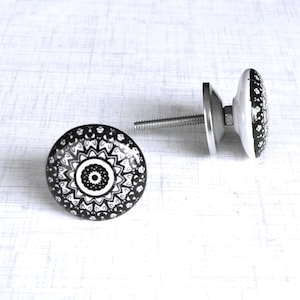 May include: Two black and white ceramic cabinet knobs with a decorative mandala design. The knobs have a round shape and a silver metal base with a threaded screw for installation. The knobs are ideal for adding a unique touch to furniture.